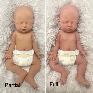 May include: Two realistic baby dolls, one with a partial paint job and the other with a full paint job, are displayed side-by-side. Both dolls are nude, wearing diapers, and lying on a fluffy, white surface. The dolls have closed eyes and detailed skin tones.