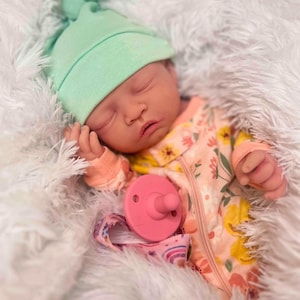 May include: A sleeping baby wearing a mint green knotted hat and a floral patterned onesie with a zipper. A pink pacifier and a rainbow-patterned pacifier clip are visible. The baby is lying on a fluffy white surface.