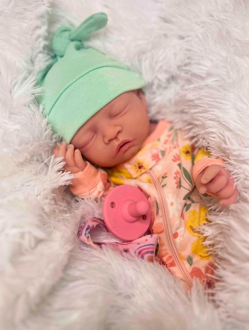 May include: A sleeping baby wearing a mint green knotted hat and a floral patterned onesie with a zipper. A pink pacifier and a rainbow-patterned pacifier clip are visible. The baby is lying on a fluffy white surface.