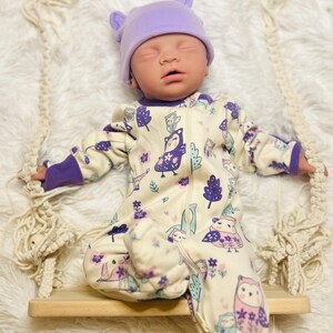 K&ouml;nnte beinhalten: Eine Babypuppe mit einem lavendelfarbenen Hut mit Ohrenakzenten und einem cremefarbenen Strampler. Der Strampler hat ein skurriles Eulen- und Blumenmuster in Lila und Rosa. Die Puppe sitzt auf einer kleinen Holzschaukel.