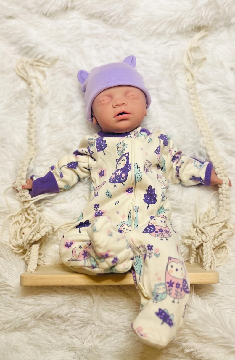 K&ouml;nnte beinhalten: Eine Babypuppe mit einem lavendelfarbenen Hut mit Ohrenakzenten und einem cremefarbenen Strampler. Der Strampler hat ein skurriles Eulen- und Blumenmuster in Lila und Rosa. Die Puppe sitzt auf einer kleinen Holzschaukel.