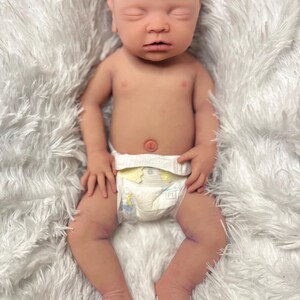 K&ouml;nnte beinhalten: Eine lebensechte Babypuppe mit geschlossenen Augen, die eine Windel tr&auml;gt. Die Puppe hat einen realistischen Hautton und liegt auf einer flauschigen wei&szlig;en Decke. Die Arme und Beine des Babys sind leicht angewinkelt.