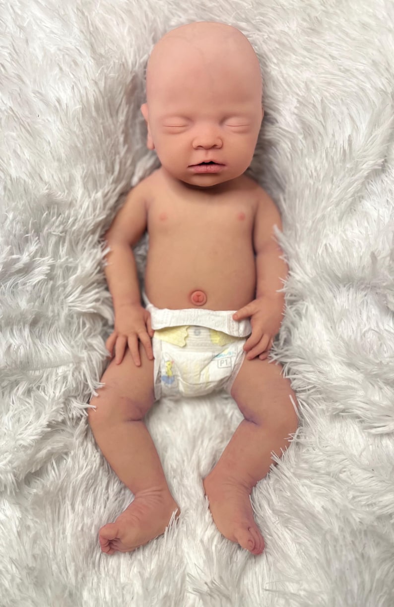 K&ouml;nnte beinhalten: Eine lebensechte Babypuppe mit geschlossenen Augen, die eine Windel tr&auml;gt. Die Puppe hat einen realistischen Hautton und liegt auf einer flauschigen wei&szlig;en Decke. Die Arme und Beine des Babys sind leicht angewinkelt.