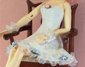 May include: A porcelain doll with pink curly hair wearing a white dress with lace trim and a pink flower brooch. The doll is sitting on a wooden chair.