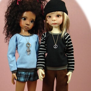 May include: Two dolls, one with brown curly hair wearing a blue sweater, plaid skirt, and pink shoes, and the other with blonde hair wearing a black and white striped sweater, brown pants, and white shoes. Both dolls are wearing necklaces.