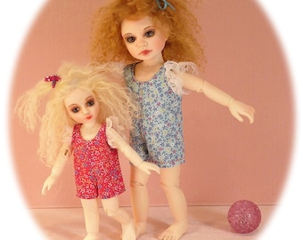 May include: Two dolls wearing floral print dresses. The doll on the left is wearing a red dress with a pink flower on her hair. The doll on the right is wearing a blue dress with a light blue bow in her hair. They are standing on a pink surface with a pink ball in the background.