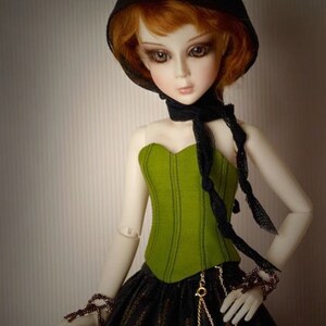 May include: A doll with red hair wearing a black hooded top, a green corset, and a black and gold skirt.