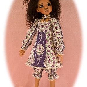 Könnte beinhalten: Eine Puppe mit braunen Locken trägt ein lila-weißes Blumenkleid mit weißer Spitzenborte und passende Blümchenhosen. Die Puppe ist barfuß und trägt eine Perlenkette mit einer lila Blume.