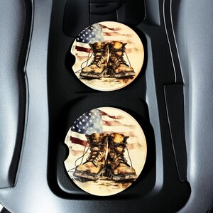 May include: Two round car coasters featuring a watercolor design of combat boots in front of a waving American flag. The coasters are tan, red, white, and blue, and are designed to fit in a car's cup holder.