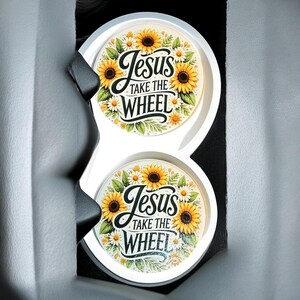 May include: Two white car coasters with a floral design featuring sunflowers and daisies. The coasters have the text "Jesus Take The Wheel" in a decorative script. The coasters are in a car cup holder.