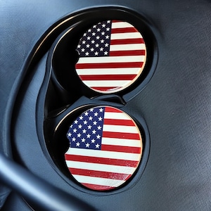 May include: Two round car coasters with the American flag design, featuring red and white stripes and a blue field of white stars. The coasters are placed in a car cup holder.