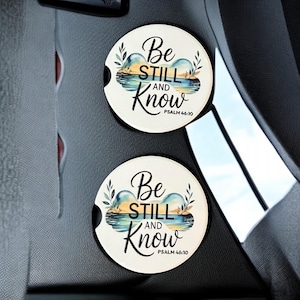 May include: Two white car coasters with the text "Be Still and Know" in black script, with a watercolor landscape design. The coasters are round and feature a slot for easy removal from the car's cup holder.