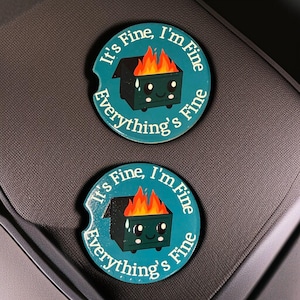 May include: Two teal car coasters with a cartoon dumpster on fire. The dumpster has a smiling face and the text "It's Fine, I'm Fine, Everything's Fine" in a circular pattern around the edge.