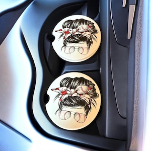 May include: Two round car coasters with a baseball theme. Each coaster features a graphic of a woman with a messy bun, sunglasses, and a baseball headband. The coasters are white with a black and red design.