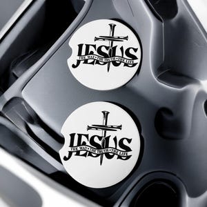 May include: Two white car coasters with the word "JESUS" in bold black letters, a cross, and a nail. The text "THE WAY THE TRUTH THE LIFE" is below the word "JESUS". The coasters are in a car cup holder.