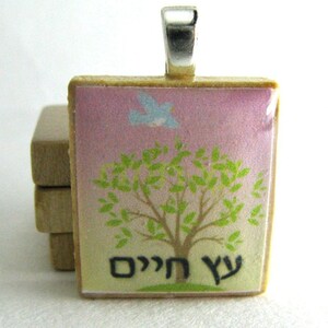 May include: A square pendant with a pink and white background featuring a small blue bird flying above a green tree. The pendant has the Hebrew words "עץ חיים" written in black at the bottom.