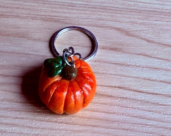 Può includere: Un piccolo portachiavi a forma di zucca arancione scolpita, caratterizzato da una foglia verde scuro e un picciolo marrone sulla parte superiore. La zucca è fissata a un anello metallico argentato, utile per agganciare chiavi o borse.