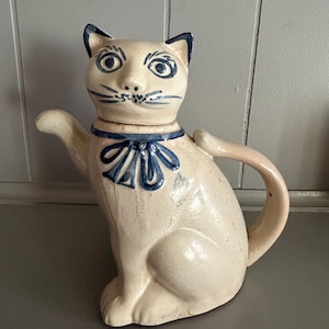 May include: A ceramic cat-shaped teapot in a seated position. The teapot is off-white with blue accents, including the ears, eyes, whiskers, and a bow around the neck. The handle and spout are light pink.
