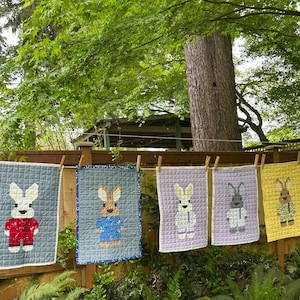 May include: Five handmade quilted wall hangings featuring different colored bunnies with different colored clothing. The bunnies are all wearing different colored clothing. The hangings are all hanging on a clothesline.