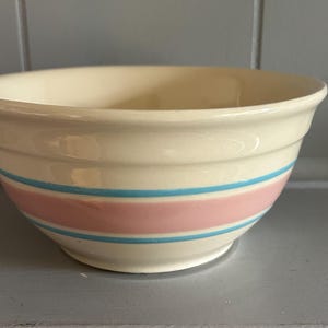 May include: A cream-colored ceramic mixing bowl featuring a wide horizontal pink band encircled by two thinner bright blue stripes. The bowl has a smooth, rounded rim and a slightly tapered base, with a glossy finish throughout the exterior surface.