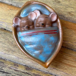 May include: A ceramic decorative piece featuring two sleeping mice nestled in a blue and red blanket within a brown, leaf-shaped dish. The mice have brown fur and pink ears. The dish has a glossy finish.