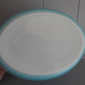 May include: An oval Pyrex baking dish with a white interior and a light blue rim. The dish has the Pyrex logo and other text printed on the bottom. Made in the USA.