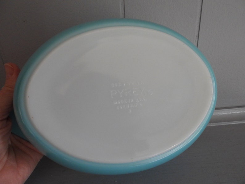 May include: An oval Pyrex baking dish with a white interior and a light blue rim. The dish has the Pyrex logo and other text printed on the bottom. Made in the USA.