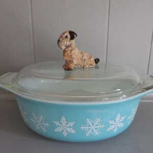 May include: A vintage turquoise Pyrex casserole dish with a clear glass lid. The dish features a white snowflake pattern and a decorative dog figurine on top. A charming addition to any kitchen.