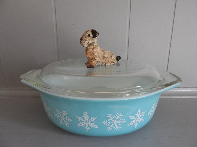 May include: A vintage turquoise Pyrex casserole dish with a clear glass lid. The dish features a white snowflake pattern and a decorative dog figurine on top. A charming addition to any kitchen.