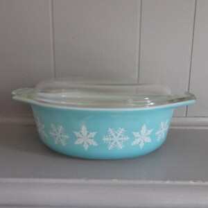 May include: A vintage turquoise Pyrex bowl with a clear glass lid. The bowl features a row of white snowflake designs around the exterior. A classic kitchenware item.