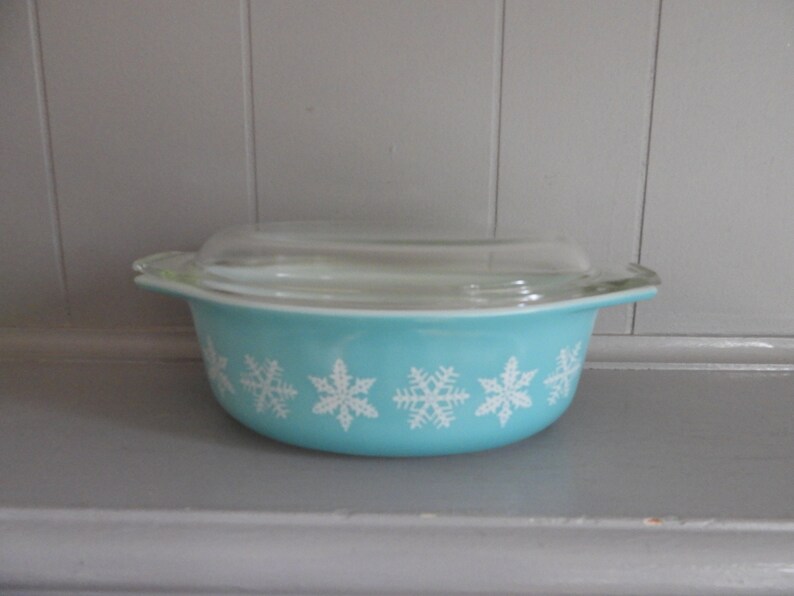 May include: A vintage turquoise Pyrex bowl with a clear glass lid. The bowl features a row of white snowflake designs around the exterior. A classic kitchenware item.