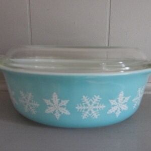 May include: A turquoise Pyrex bowl with a clear glass lid. The bowl features a white snowflake pattern around the exterior. The bowl has two handles and is designed for oven use. A classic kitchenware item.