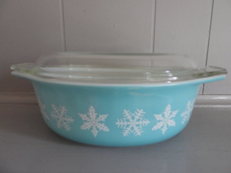 May include: A turquoise Pyrex bowl with a clear glass lid. The bowl features a white snowflake pattern around the exterior. The bowl has two handles and is designed for oven use. A classic kitchenware item.