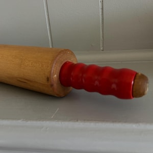 May include: A wooden rolling pin with a red handle. The rolling pin is made of light-colored wood and has a smooth, cylindrical body. The handle is a vibrant red color and has a ridged design for grip. Kitchenware.