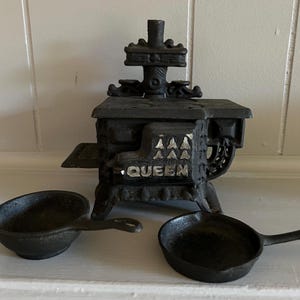 May include: A miniature black cast iron stove with the word "QUEEN" on the front, accompanied by two small cast iron skillets. The stove has intricate detailing and a chimney. The items are displayed on a white surface.