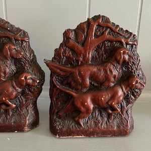 May include: Pair of vintage brown bookends featuring detailed relief carvings of dogs in a forest setting. The bookends have a rustic, aged appearance, with visible texture and color variations. The dogs are depicted in a dynamic pose, adding visual interest.