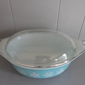 May include: A vintage Pyrex casserole dish with a clear glass lid. The bowl is light blue with white snowflake designs around the base. The lid has a clear, rounded shape. This dish is ideal for baking and serving.
