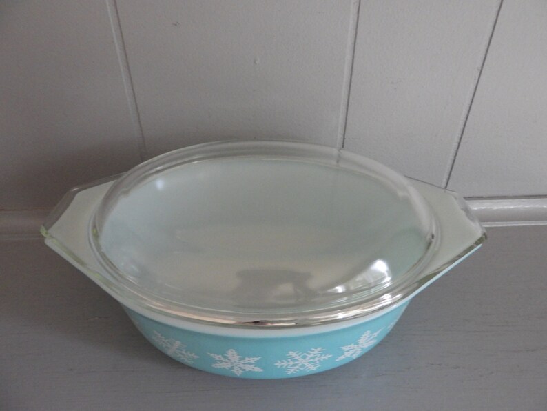 May include: A vintage Pyrex casserole dish with a clear glass lid. The bowl is light blue with white snowflake designs around the base. The lid has a clear, rounded shape. This dish is ideal for baking and serving.