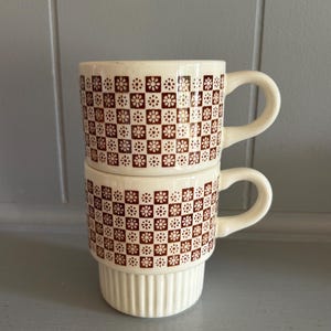 May include: Two stacked cream-colored ceramic mugs featuring a repeating brown grid pattern with small floral motifs inside each square. The mugs have a smooth upper section with the pattern and a vertically ribbed base, each with a curved handle.