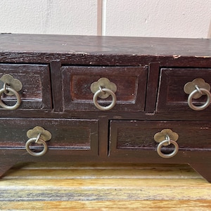 May include: A dark brown wooden jewelry box with five drawers. Each drawer has a decorative metal pull with a ring handle. The corners are reinforced with metal brackets. The box sits on a light-colored surface.