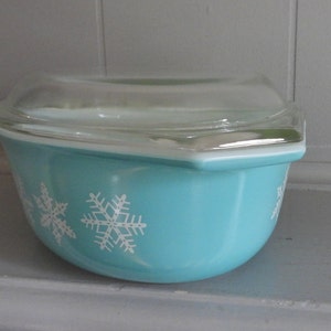 May include: A turquoise Pyrex bowl with a clear glass lid. The bowl features white snowflake designs. The bowl is a classic kitchenware item, perfect for serving or storing food.