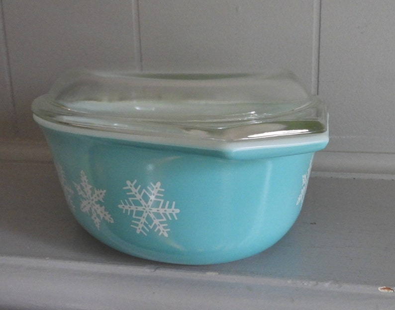 May include: A turquoise Pyrex bowl with a clear glass lid. The bowl features white snowflake designs. The bowl is a classic kitchenware item, perfect for serving or storing food.