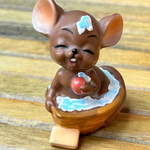 May include: A brown ceramic mouse figurine with closed eyes, holding a red apple. The mouse is in a brown boat with blue and white accents, and a small cheese block is in front of the boat. The figurine is on a wooden surface.