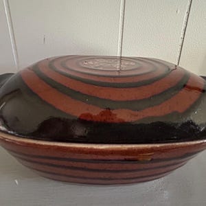 May include: A ceramic serving dish with a lid. The dish is oval-shaped and features a glossy finish. The lid and base are decorated with concentric bands of brown and black. The dish has two small handles on either side.