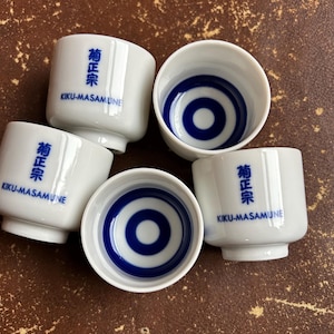 May include: Five white ceramic sake cups with blue concentric circle designs inside. Each cup has the text "菊正宗 KIKU-MASAMUNE" printed in blue. The cups are arranged on a brown surface.