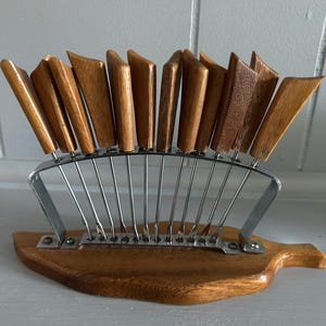 May include: A vintage wooden cheese knife set with a metal and wood stand. The knives have angled wooden handles and are arranged in a fan shape. The base is a wooden cutting board shape.