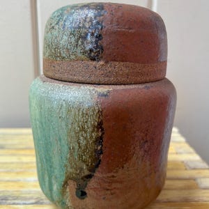 May include: A cylindrical ceramic jar with a matching lid, featuring a split color design of earthy terracotta red and muted sage green. The surface has a textured, speckled finish with a dark, uneven vertical line separating the two colors.