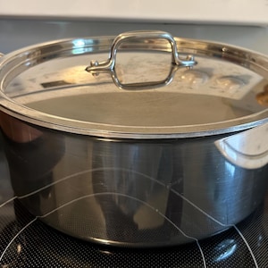 May include: A stainless steel cooking pot with a matching lid. The pot has two handles and a handle on the lid. The pot is sitting on a black stovetop. The pot is made of shiny metal.