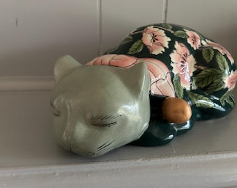 May include: A ceramic cat figurine in a sleeping pose. The cat's head is a pale green, while its body is covered in a dark green floral pattern with pink flowers and green leaves. A small, gold-colored ball is near the cat.