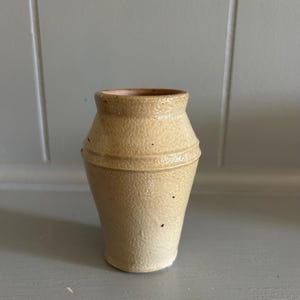 May include: A small, beige ceramic vase with a textured, salt-glazed finish. The vessel features a tapered neck, a slightly flared rim, and a distinct horizontal ridge encircling the upper body, showcasing a natural, earthy appearance.